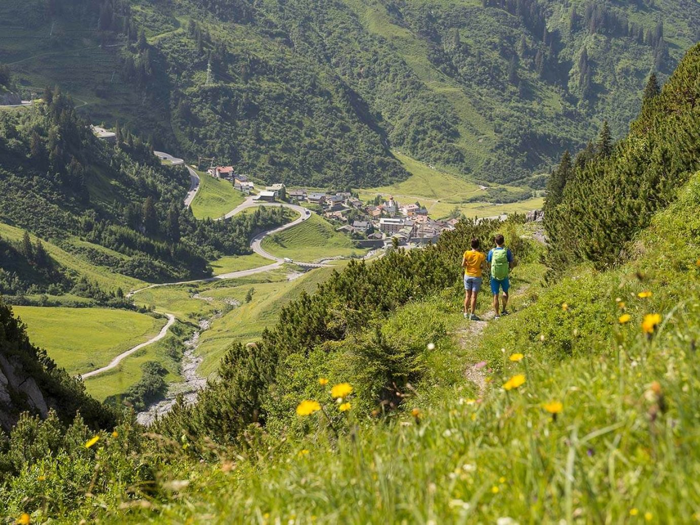 2 Personen mitten in den Bergen, rundum grüne Gegend mit Wald und Ortschaft Stuben am Arlberg im Hintergrund zu sehen.